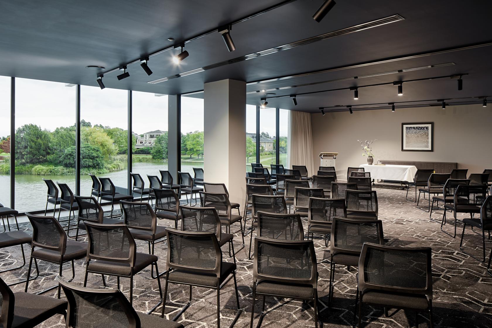 A hotel event space with floor-to-ceiling windows overlooking a lake and rows of black chairs at Punthill Caroline Springs.