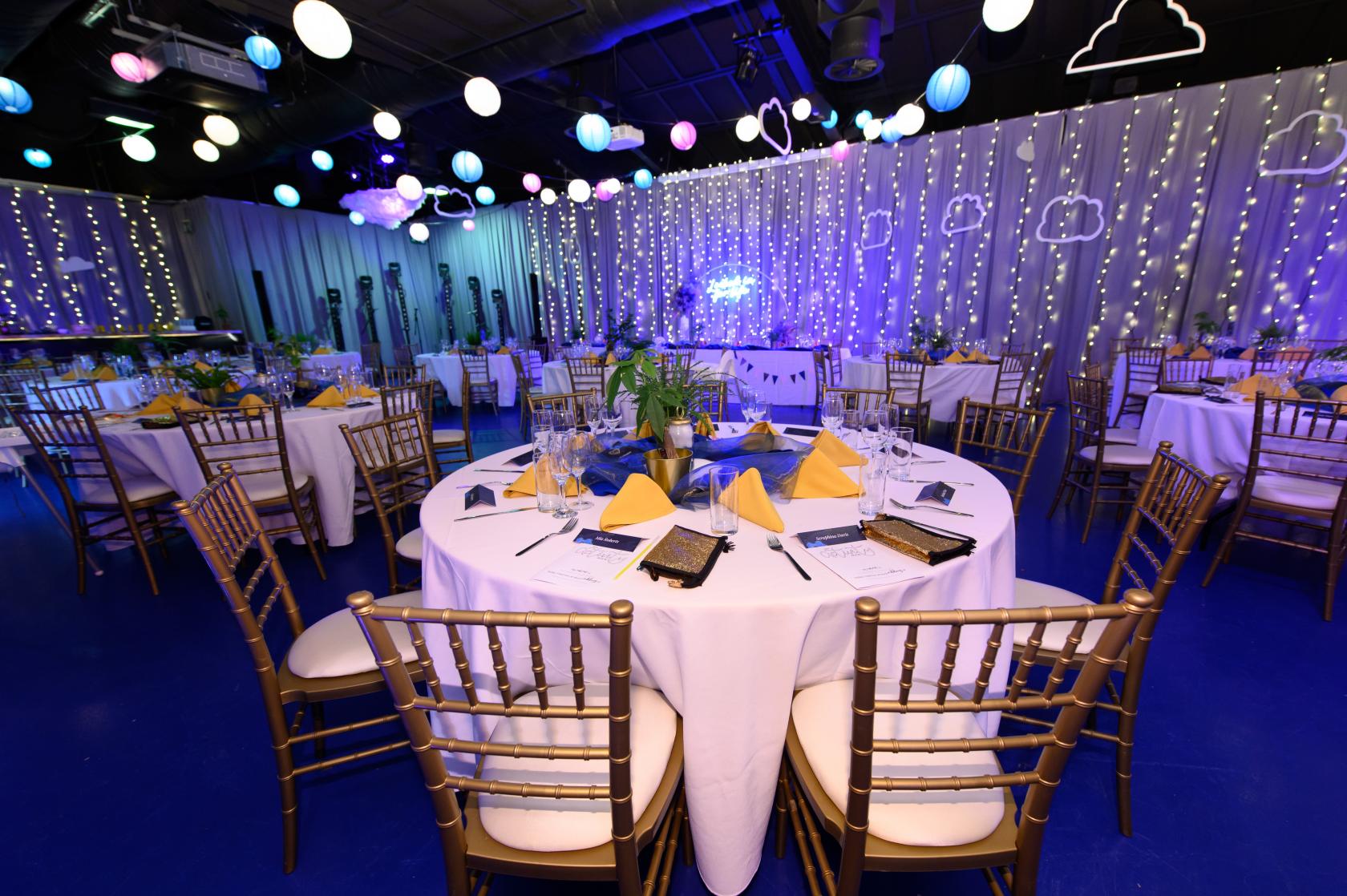 An event space with round tables set with yellow napkins, string lights, and hanging paper clouds at The Crystal Maze LIVE Experience.