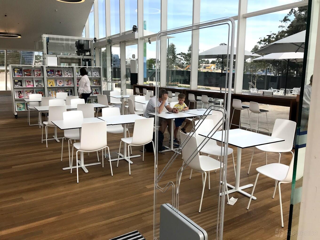 A cafe with floor-to-ceiling windows, white chairs, and wooden flooring at Noun Green Square.