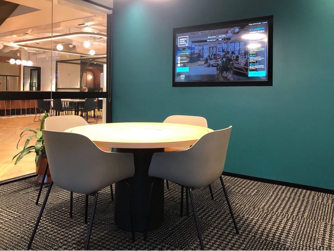 A coworking meeting room with a round table, four modern chairs, teal walls, and a wall-mounted screen at JustCo at Collins St.
