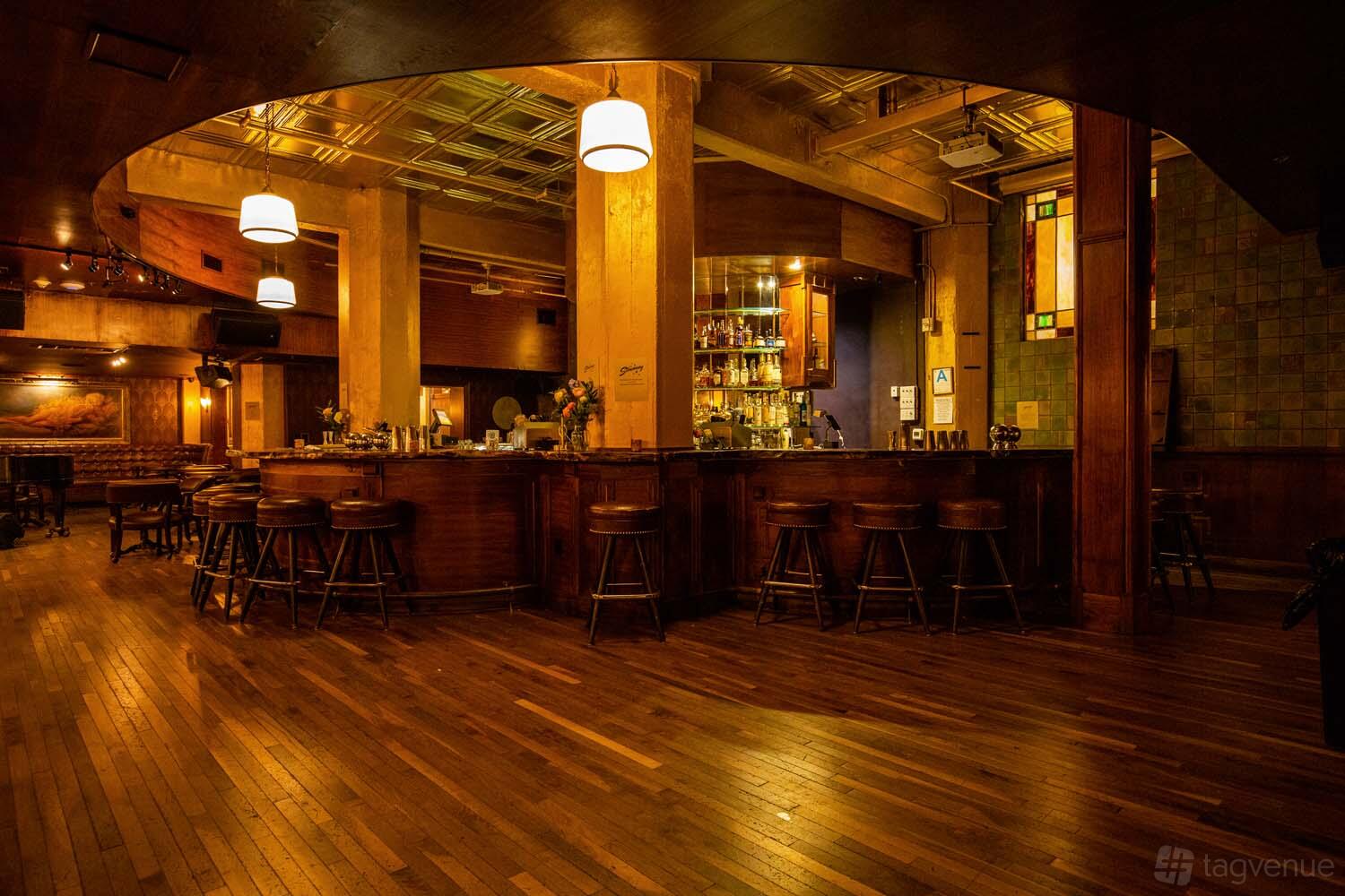 A bar with wooden floors, high stools, brass fixtures, and warm ambient lighting at The Stowaway DTLA.