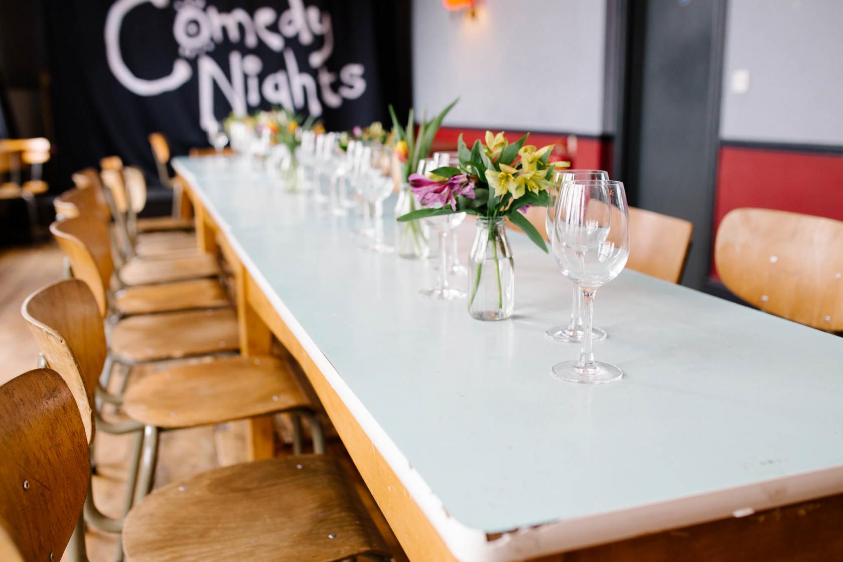 A dining room with a long light blue table, wooden chairs, glassware, and flower vases at The Honor Oak.