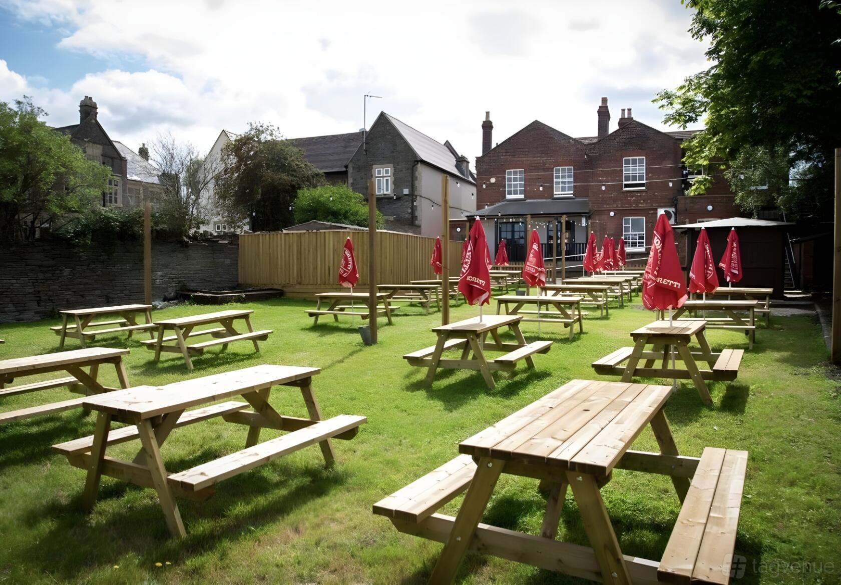 An outdoor pub space with wooden picnic tables and red umbrellas on a grassy lawn at Fishponds Tap.