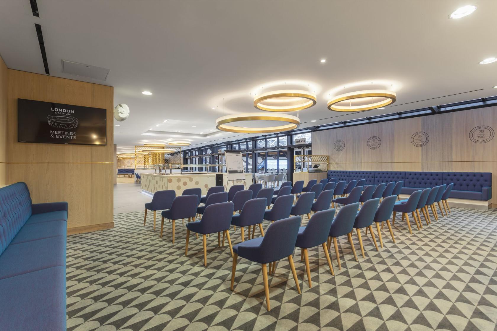 An auditorium with rows of blue chairs, geometric carpet, and circular pendant lights at London Stadium's The Royal East.