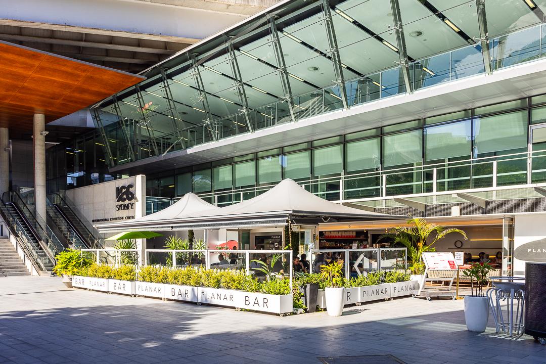 An outdoor restaurant space with a canopy, glass fencing, and greenery at Planar Restaurant.
