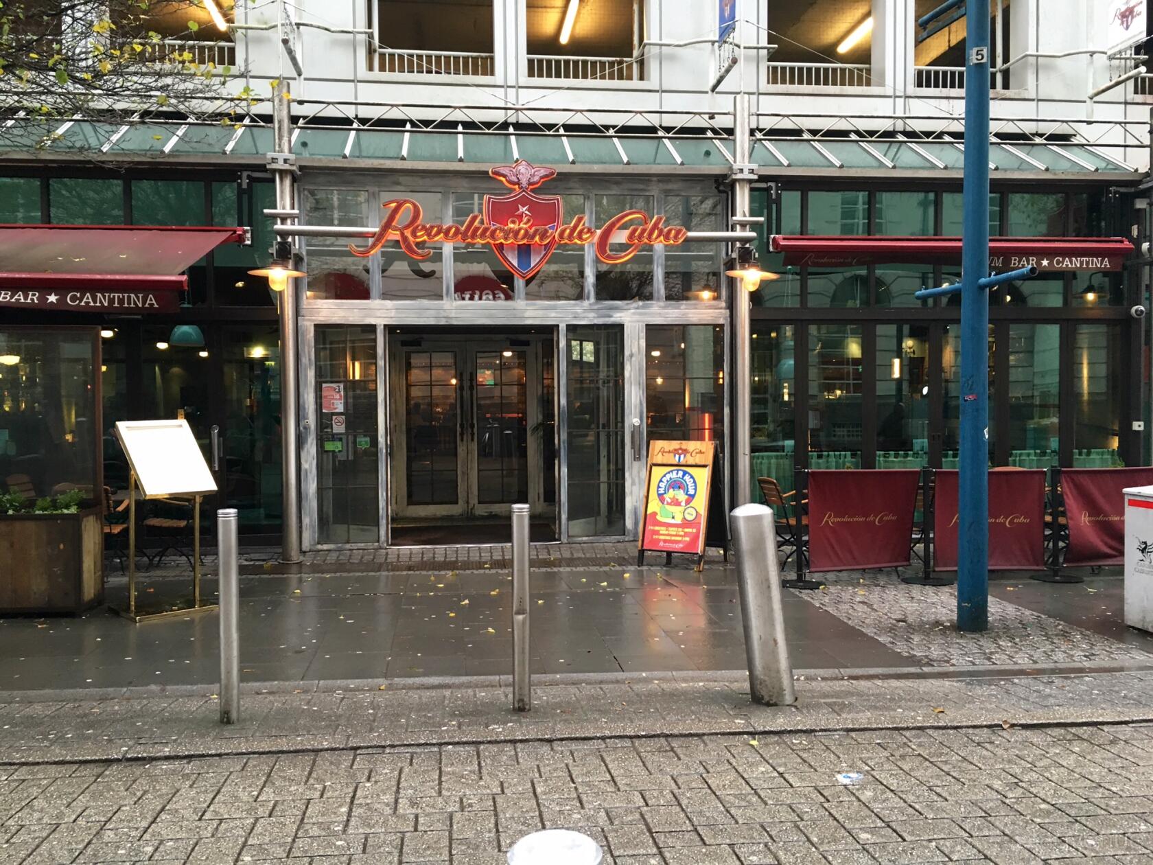 The exterior of a pub with glass doors, red signage, covered outdoor seating and sidewalk at Revolucion De Cuba Cardiff.