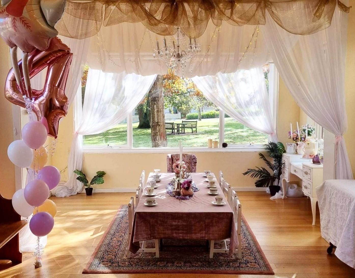 A function room with sheer curtains, a long table set for tea, pastel balloons, and garden views at True Fairies.