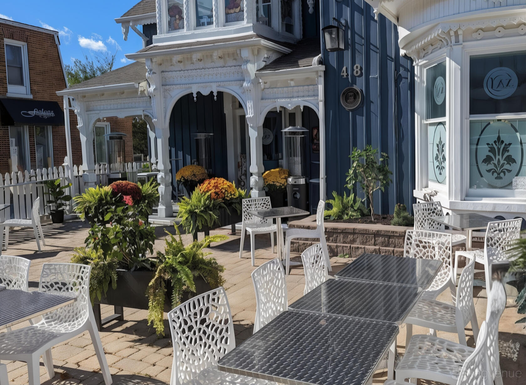 An outdoor restaurant patio with metal tables, white chairs, potted plants, and Victorian architecture at Laz Authentic Cuisine Inc.