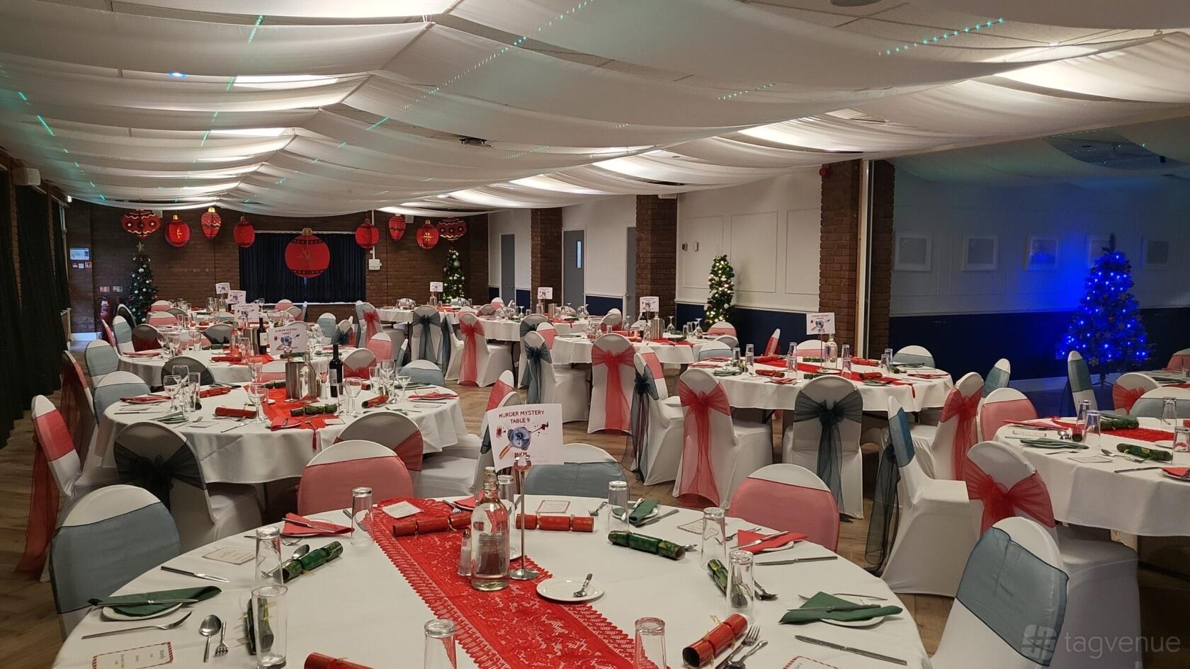 An event venue with round tables draped in white linens, red accents, and lantern decorations at Newbury Rugby Club.