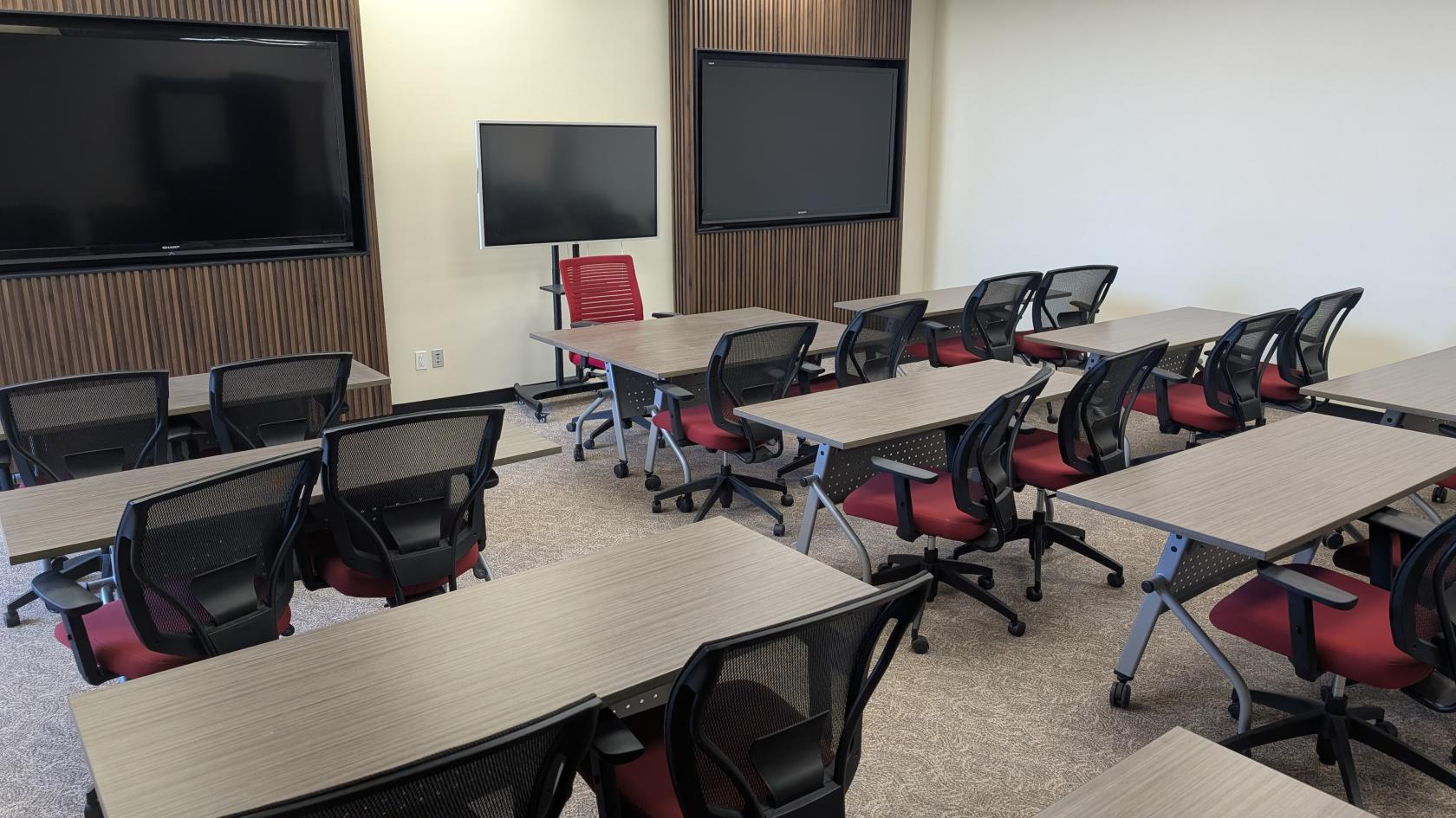 A training room with rows of tables, mesh chairs with red seats, and two wall-mounted screens at Learner First Studio.