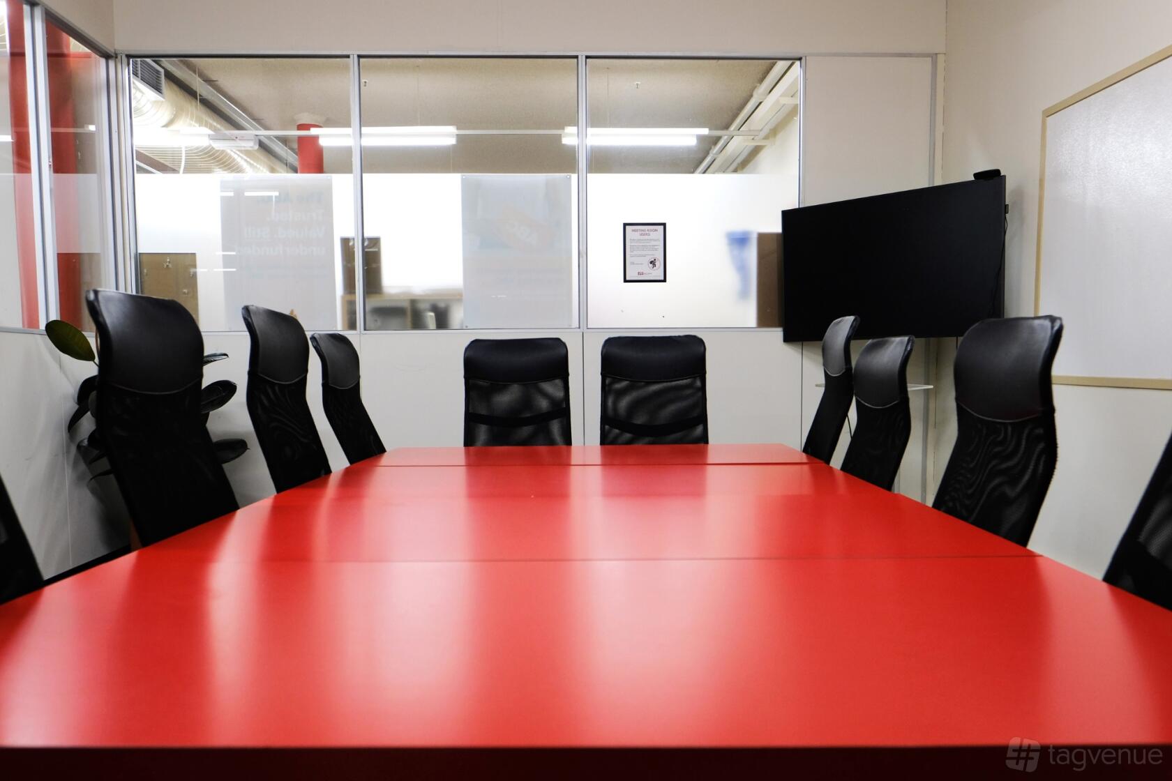 A meeting room with a long red conference table, black chairs, glass wall, and wall-mounted TV at Ross House.