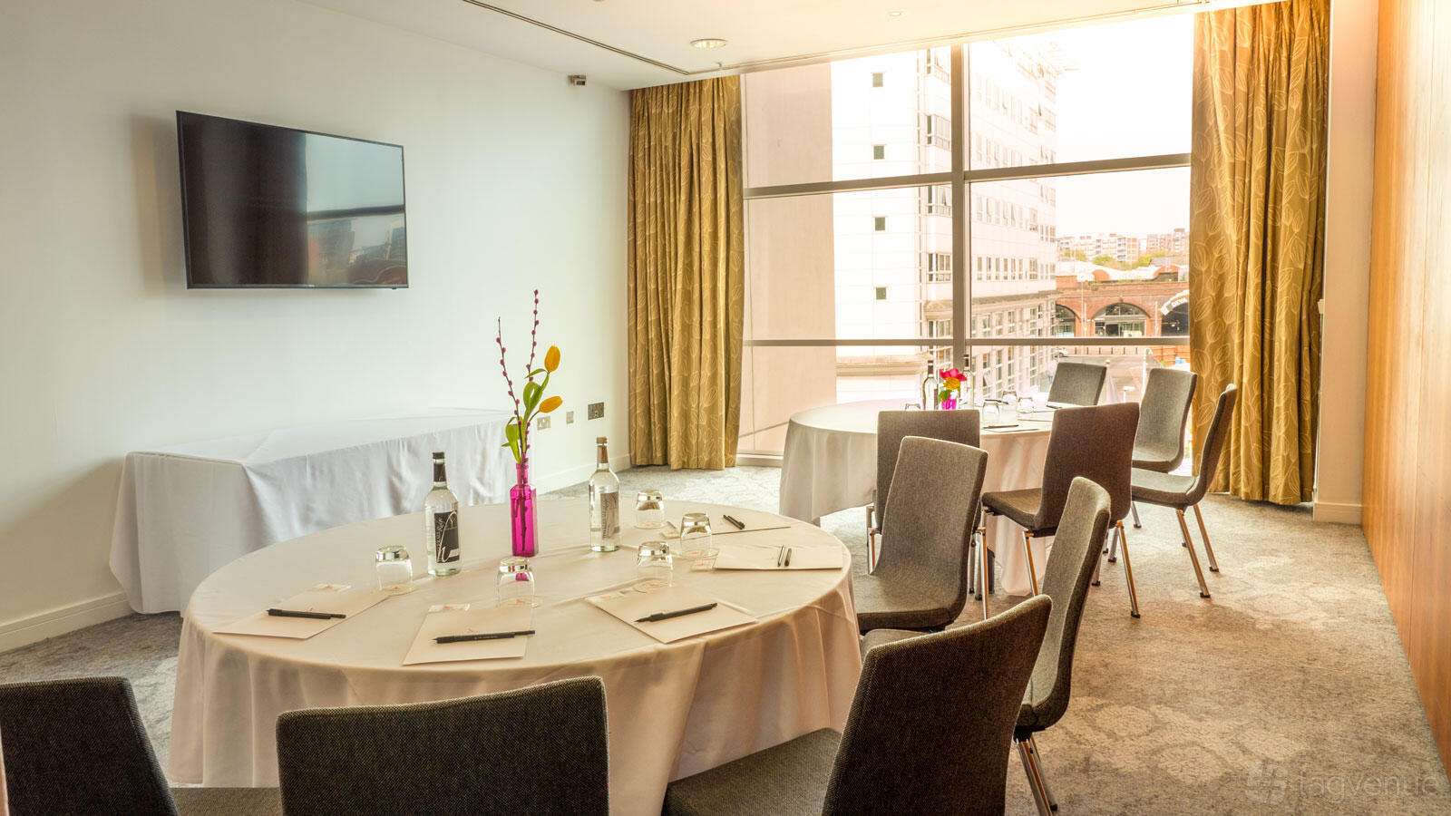 A hotel meeting room with round tables, bottled water, notepads, and floor-to-ceiling windows at The Lowry Hotel.