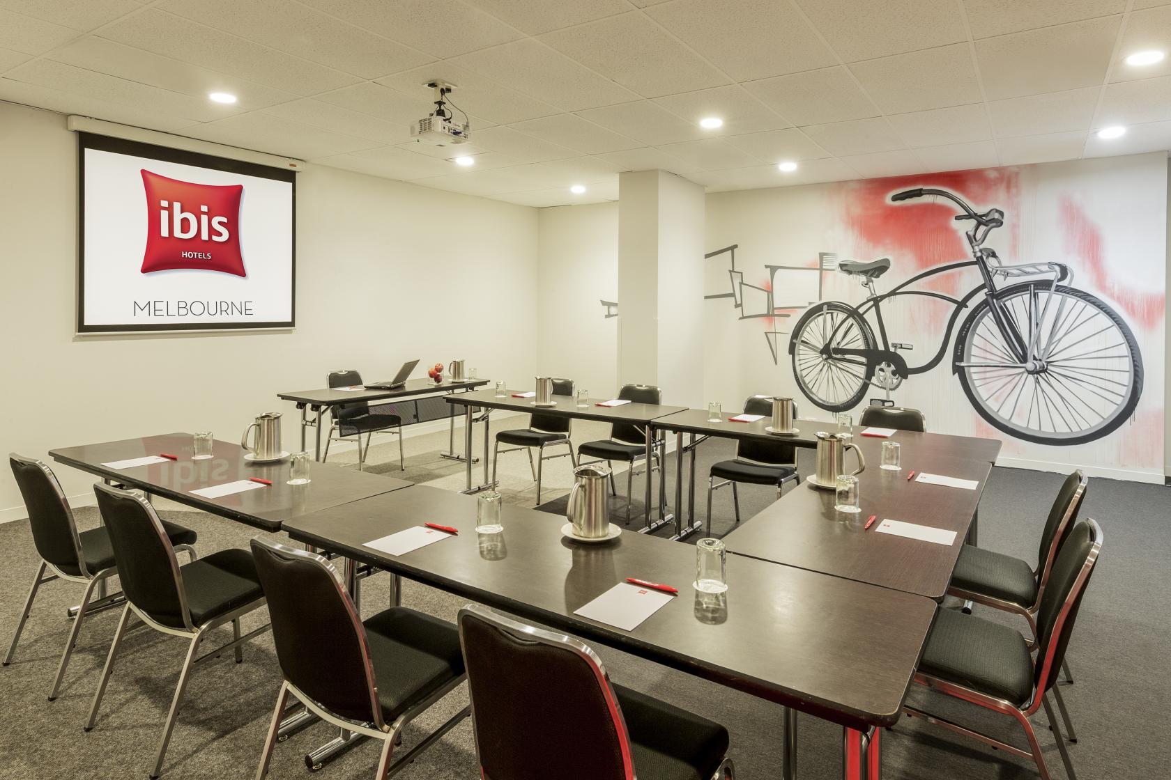 An event space in a hotel with U-shaped tables, chairs, a projector screen, and a bicycle mural at Ibis Melbourne Hotel and Apartments.