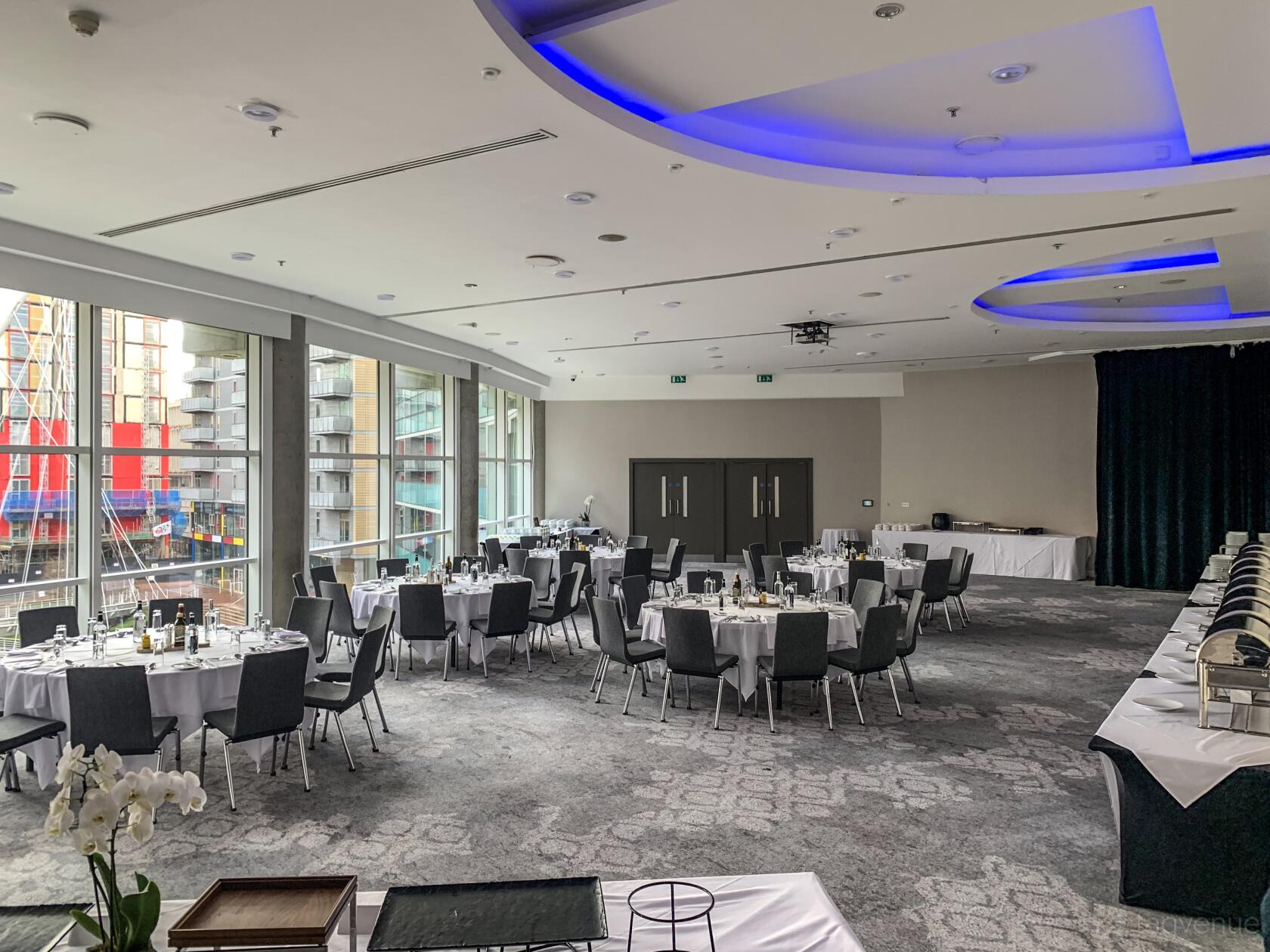 A hotel ballroom with floor-to-ceiling windows, round tables with white linens, and blue ceiling accent lights at The Lowry Hotel.