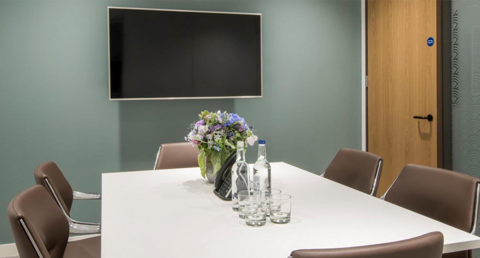 A meeting room with a large wall-mounted screen, floral centerpiece, and brown chairs at 1 King William Street.