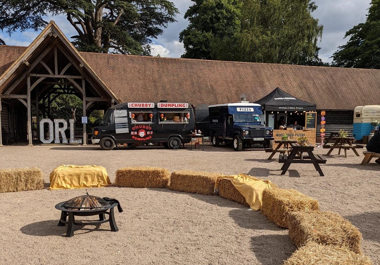 An outdoor barn space with food trucks, picnic tables, hay bales, and a fire pit at Knebworth House & Barns.