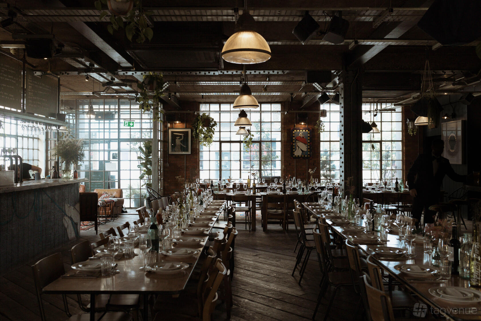 A bar with long banquet tables set for dining, wooden floors, and large industrial windows at Never For Ever.