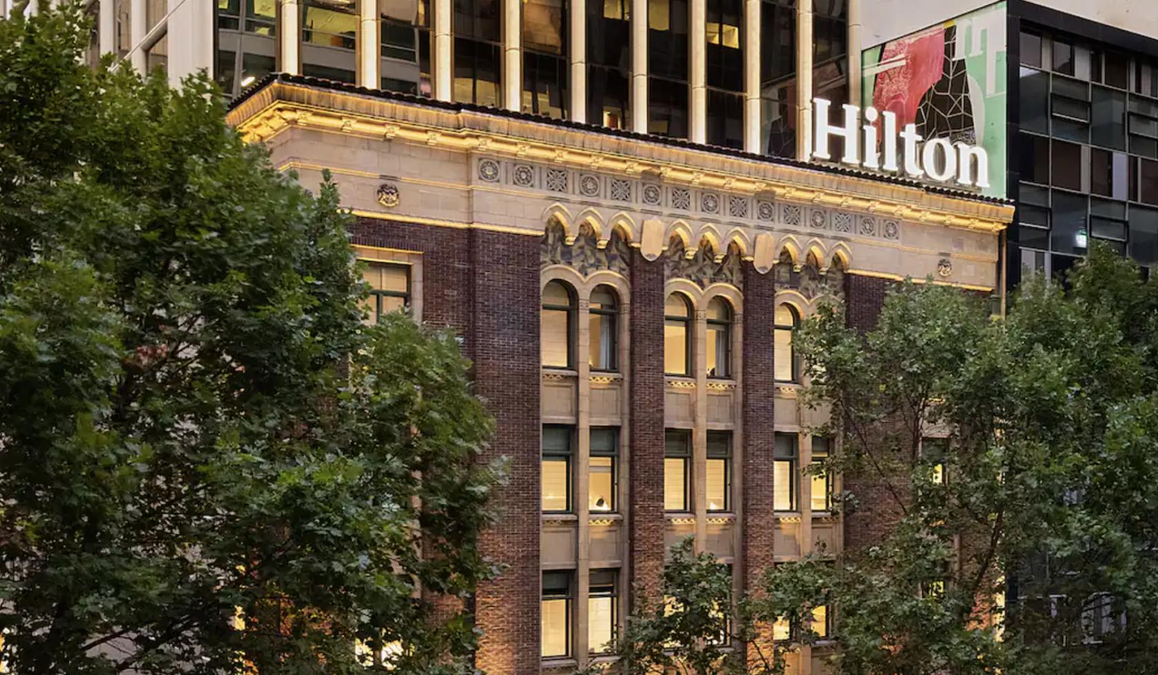 A hotel with a historic brick facade, arched windows, and illuminated architectural details at Hilton Melbourne Little Queen Street.