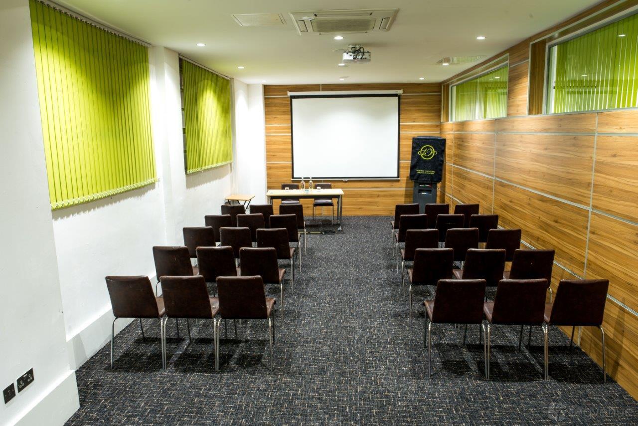 A meeting room with rows of black chairs facing a projection screen and wood-paneled walls at The Wesley Euston Hotel.