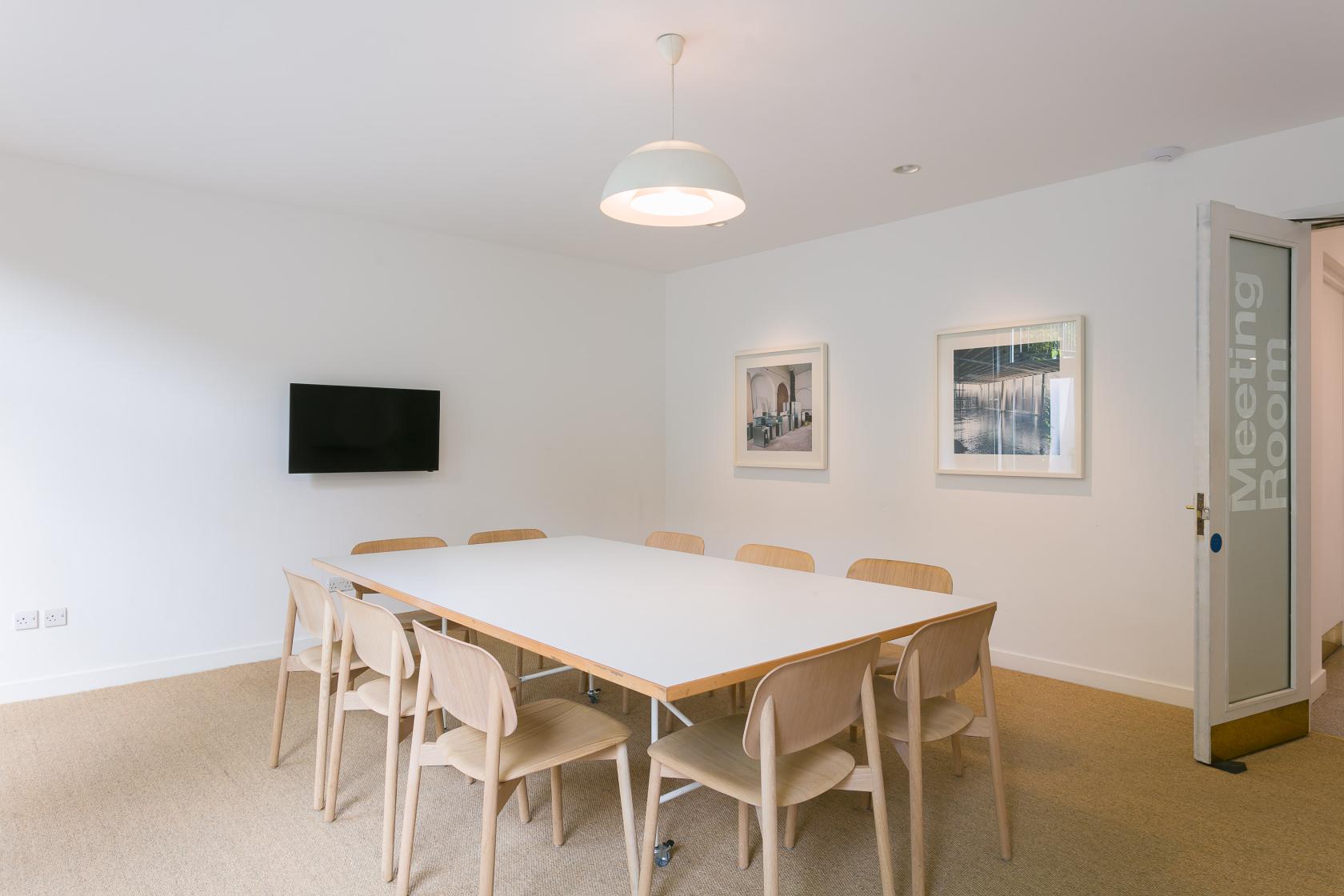 A meeting room with a large white table, light wooden chairs, wall-mounted TV, and framed art at Camden Art Centre.