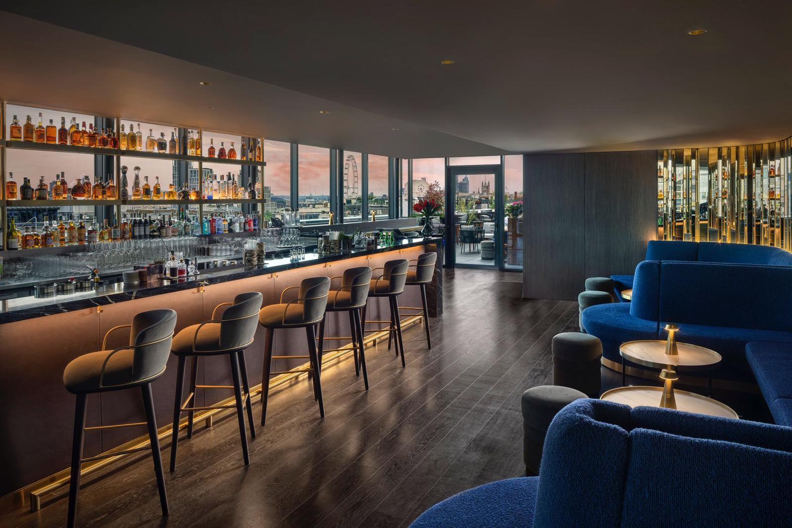A rooftop bar with floor-to-ceiling windows, backlit shelves with bottles, and plush blue seating at AMANO Hotel Covent Garden.