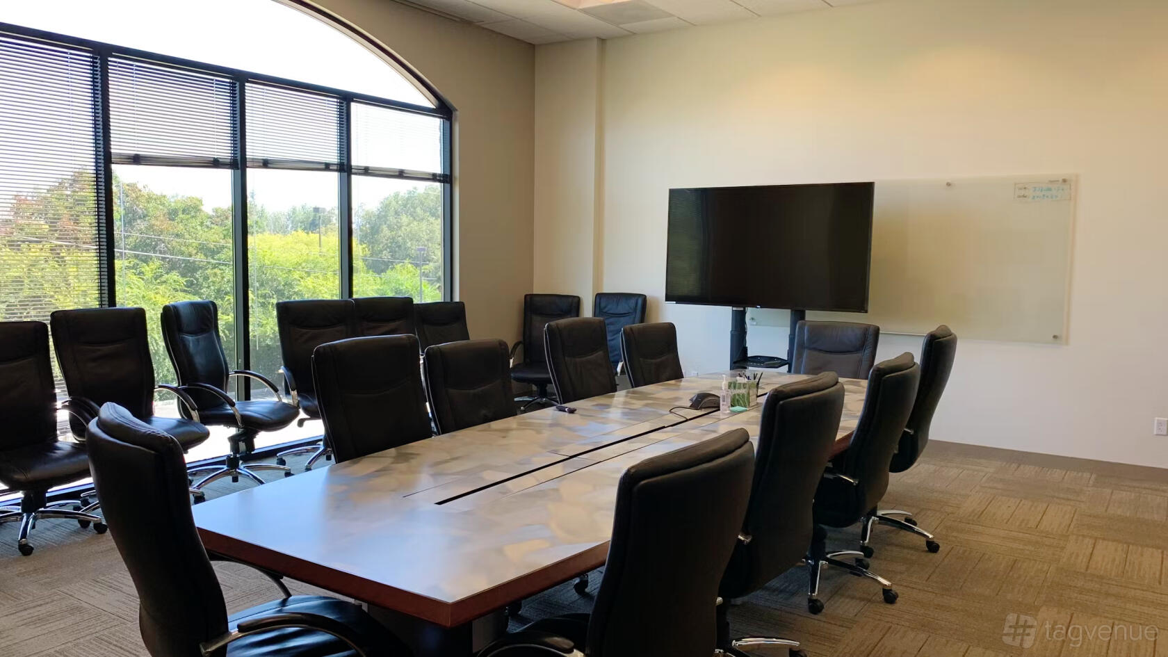 A coworking conference room with a long table, black leather chairs, floor-to-ceiling windows, and a large screen at JJ Lake Business Center.