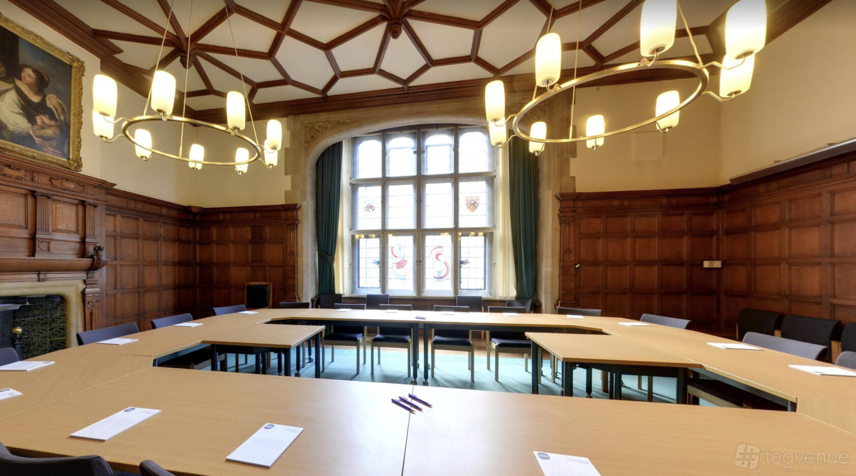 A hall with wood-paneled walls, a large stained glass window, and U-shaped tables beneath chandeliers at Oxford Town Hall.