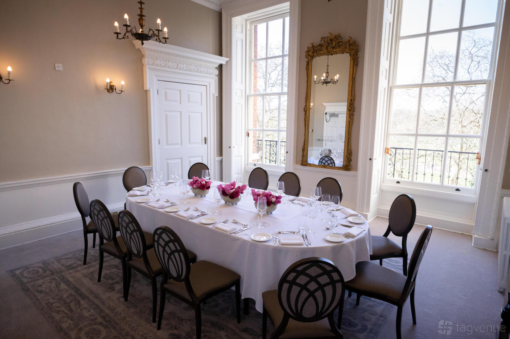 A dining room with tall windows, a large ornate mirror, chandelier lighting, and an oval table with chairs at Six Park Place.