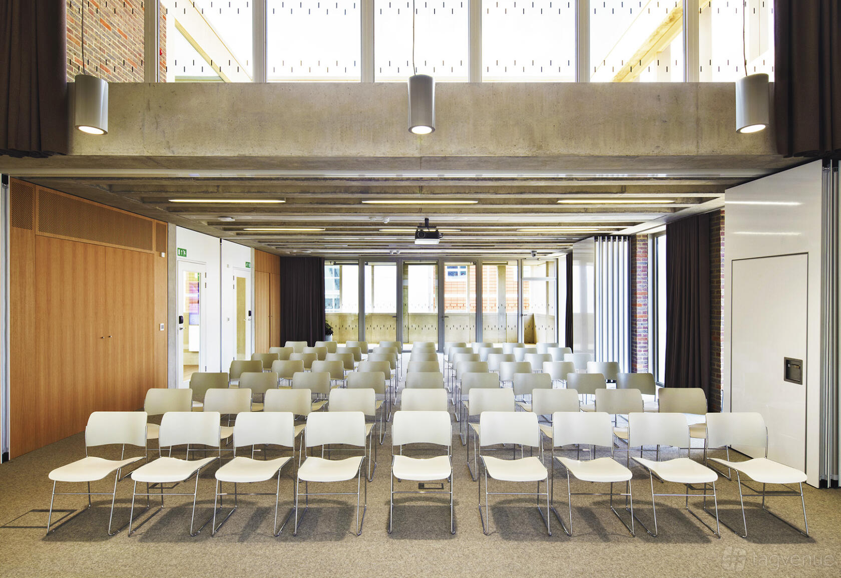 A function room with rows of white chairs, exposed concrete beams, and floor-to-ceiling windows at ORTUS Conference and Events Venue.