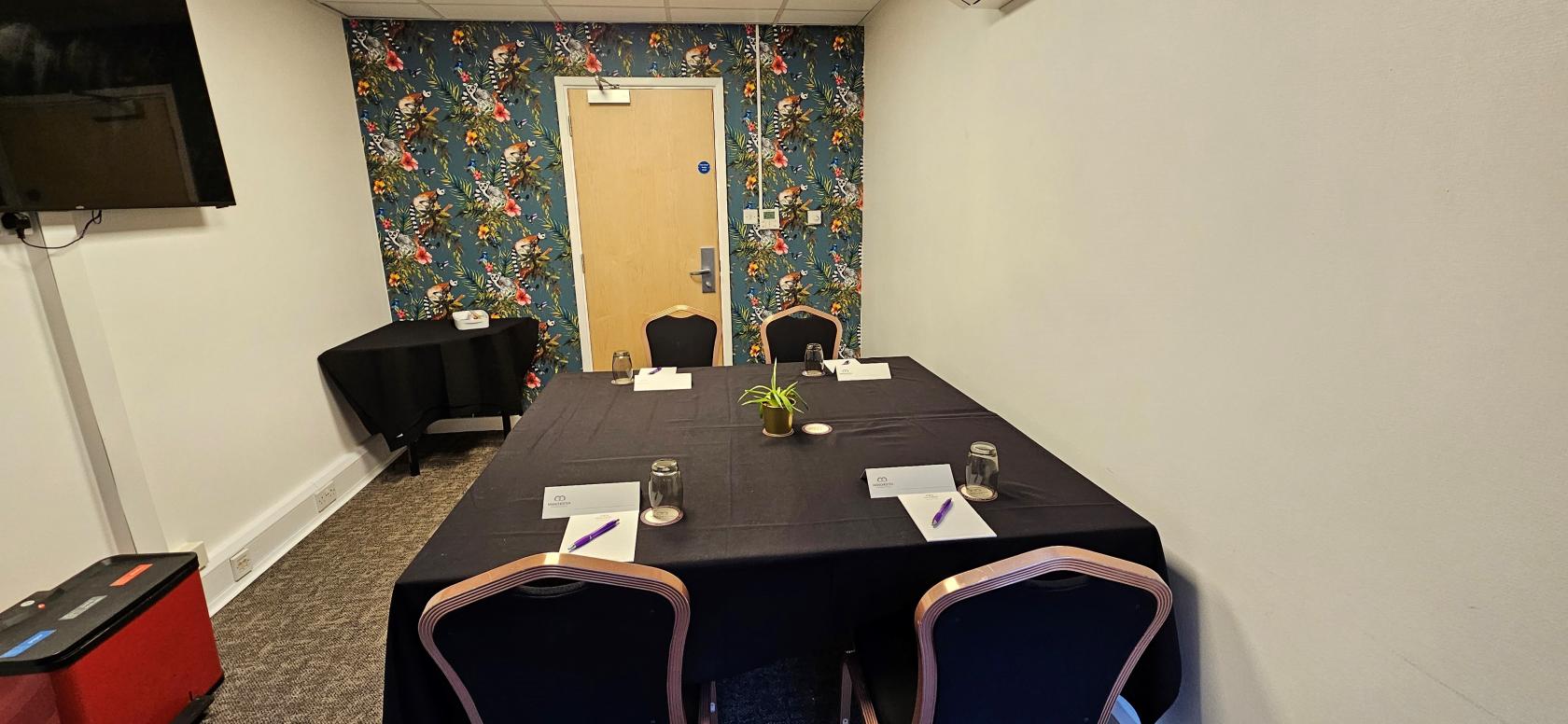 A hotel meeting room with a floral accent wall, rectangular table, notepads, and black tablecloths at Manchester Conference Centre & Pendulum Hotel.