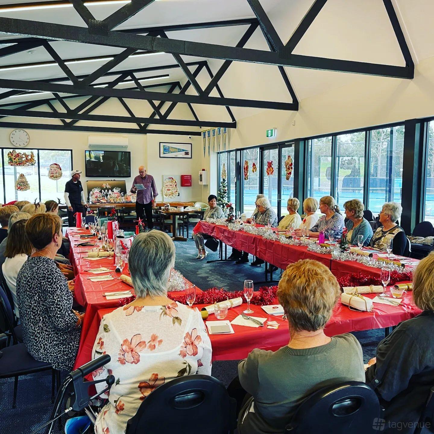 An event venue with long tables covered in red tablecloths, decorated beams, and floor-to-ceiling windows at Frankston Centenary Tennis Club.