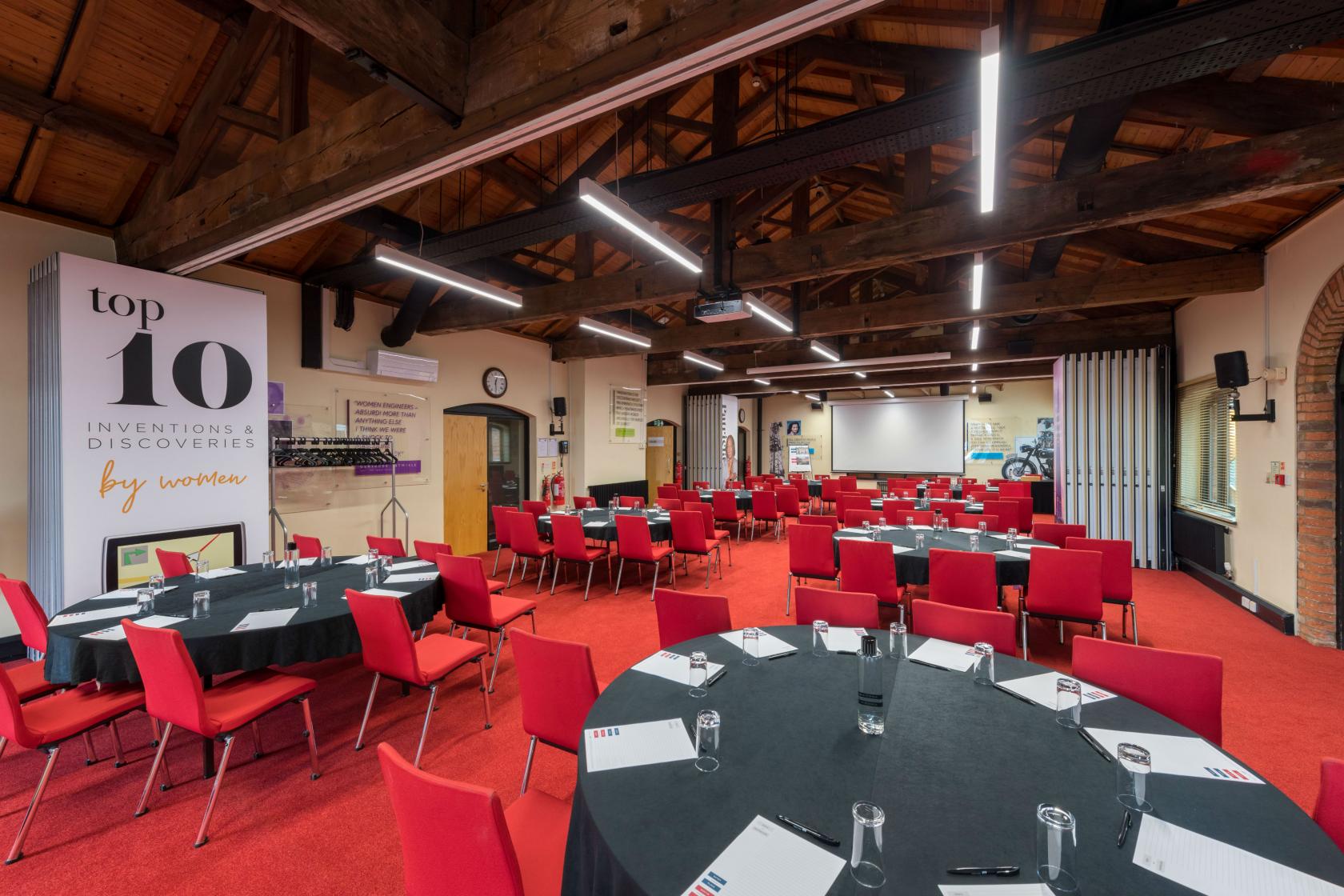A function room with exposed wooden beams, red chairs, and round tables set for meetings at IET Birmingham: Austin Court.