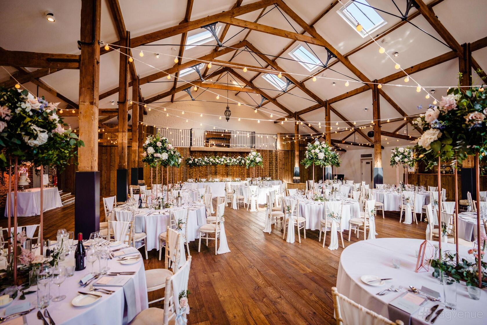 A hall with exposed wooden beams, string lights, and round tables with tall floral centerpieces at Oxnead Hall.