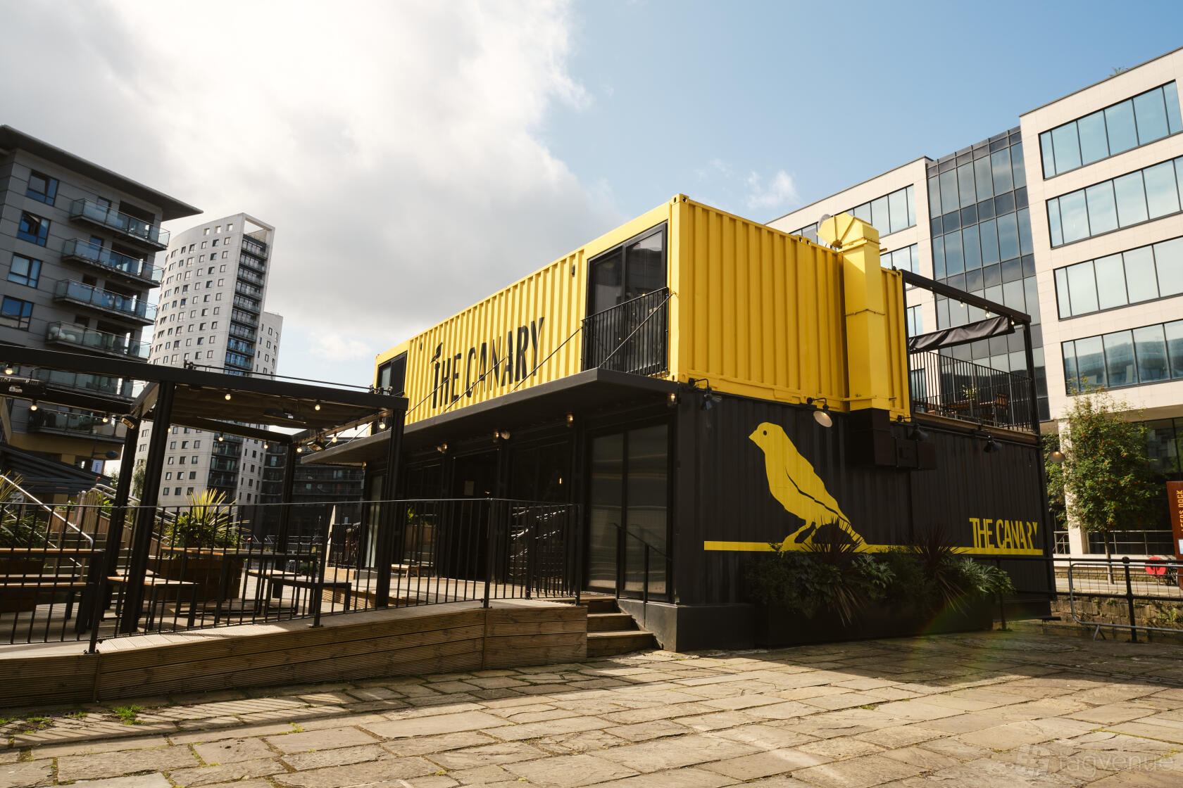 A bar in a converted shipping container with a yellow and black facade and outdoor seating at Canary.