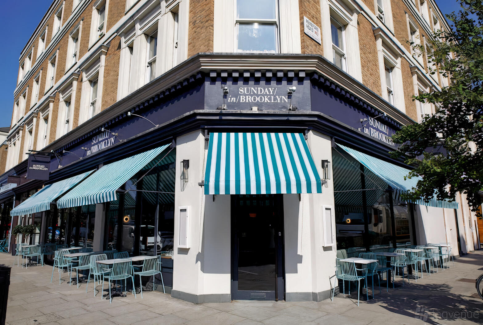 A corner restaurant with blue and white striped awnings and outdoor seating at Sunday in Brooklyn Notting Hill.