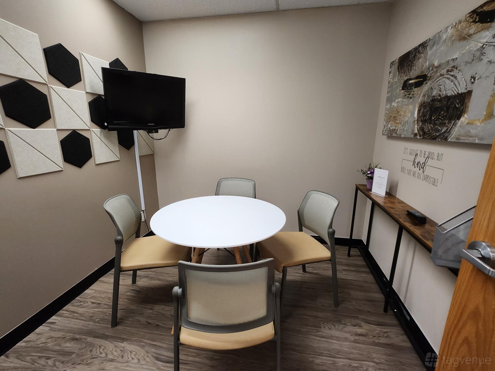 A meeting room with a round table, four chairs, wall-mounted TV, geometric wall panels, and artwork at AHANSA Business Center.