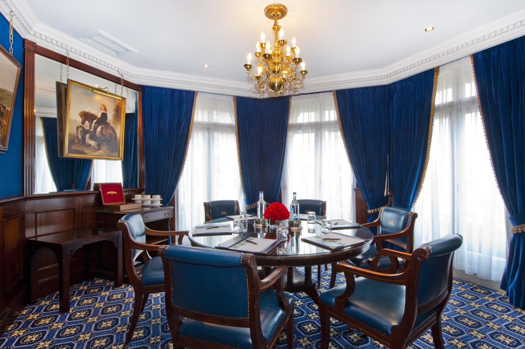 A hotel dining room with a round wooden table, blue leather chairs, chandelier, and floor-to-ceiling curtains at The Chesterfield Mayfair.