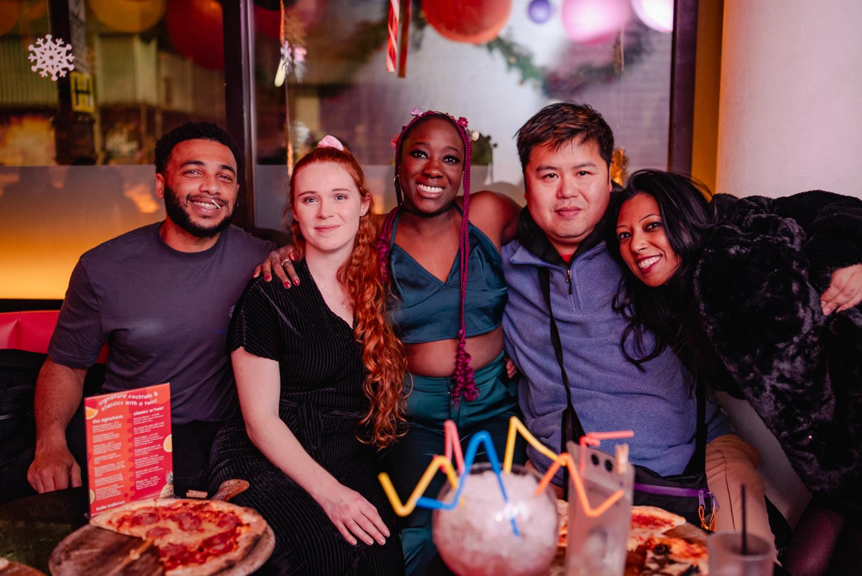 A cocktail bar with pink neon lighting, colorful hanging decorations, and pizza on tables at Ballie Ballerson London.