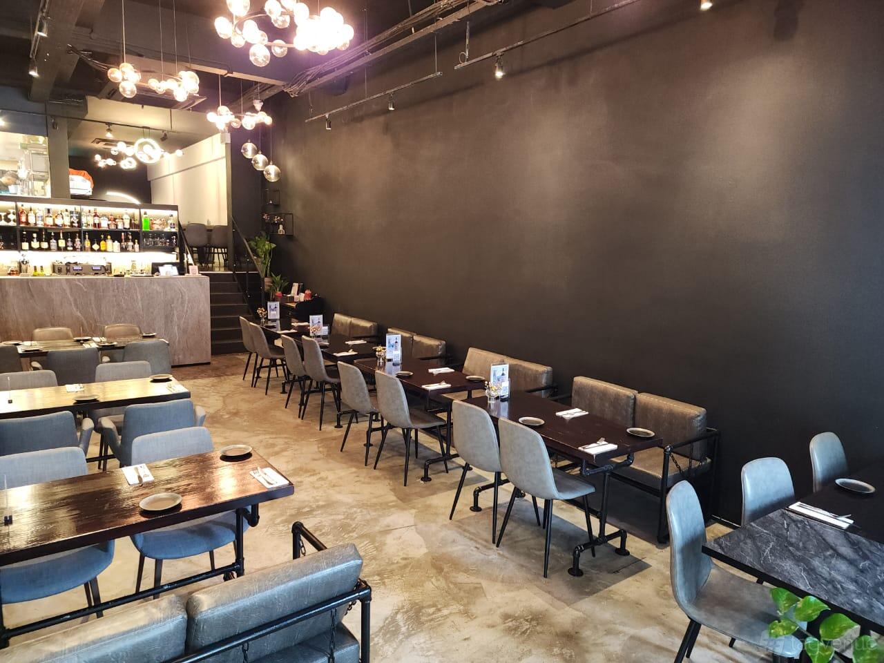 A restaurant with dark wooden tables, gray cushioned chairs, exposed lighting, and a marble bar at Perle Noire Oyster & Grill Bar.