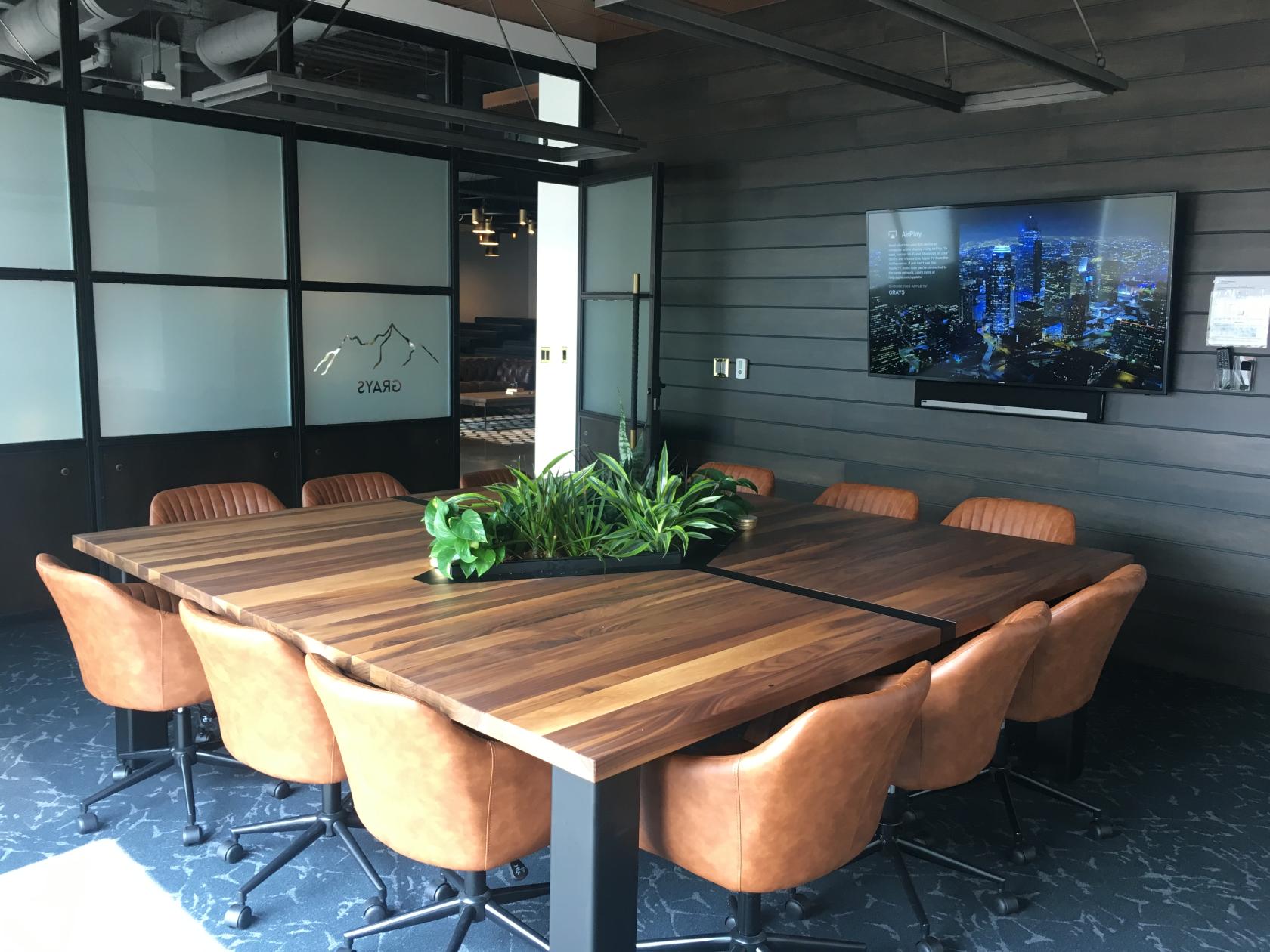 A coworking meeting room with a large wooden conference table, leather chairs, and a wall-mounted screen at Venture X LoDo.