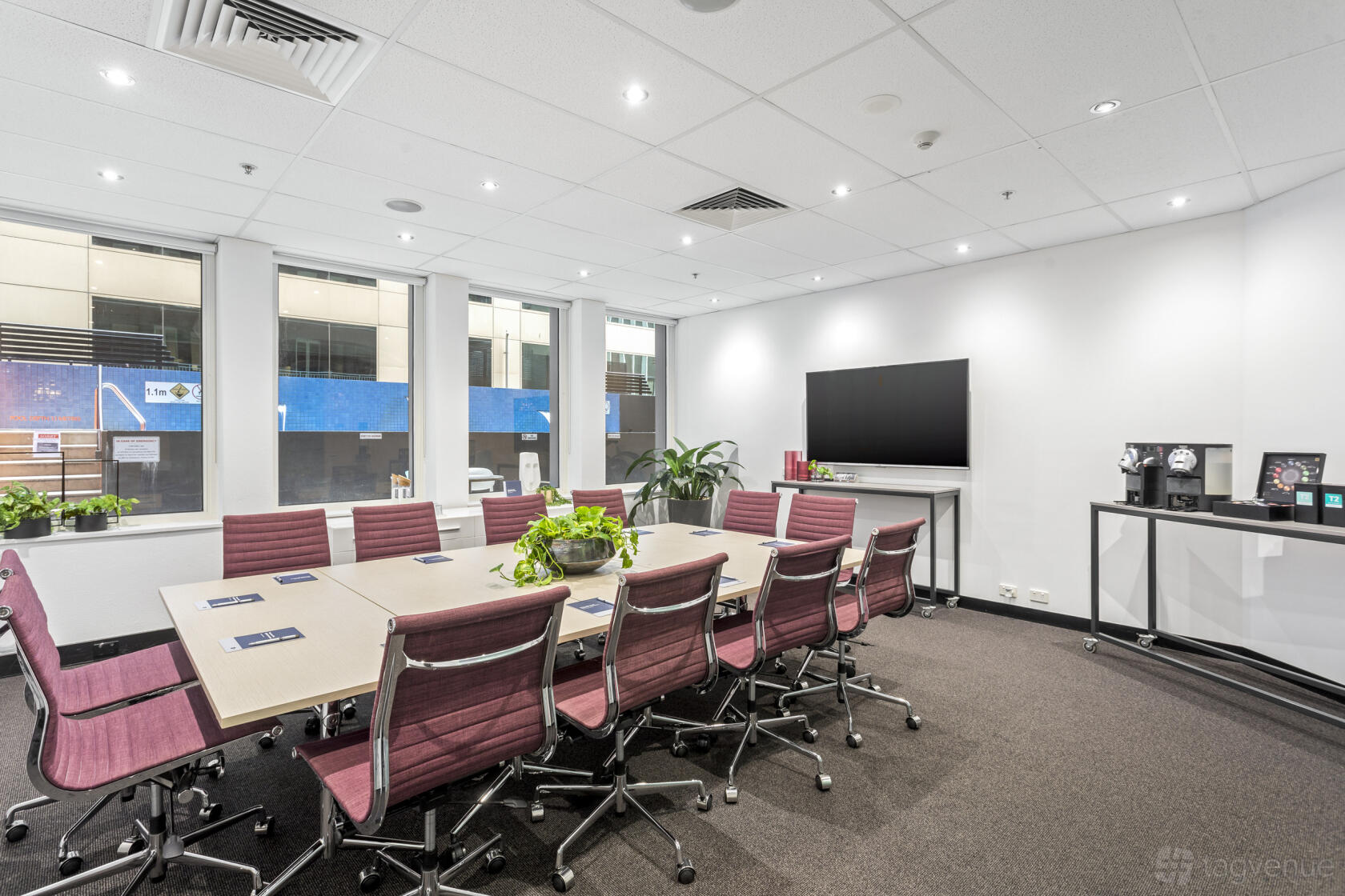 A boardroom with burgundy chairs, rectangular tables, large windows, and a wall-mounted TV at Collins Street Tower.
