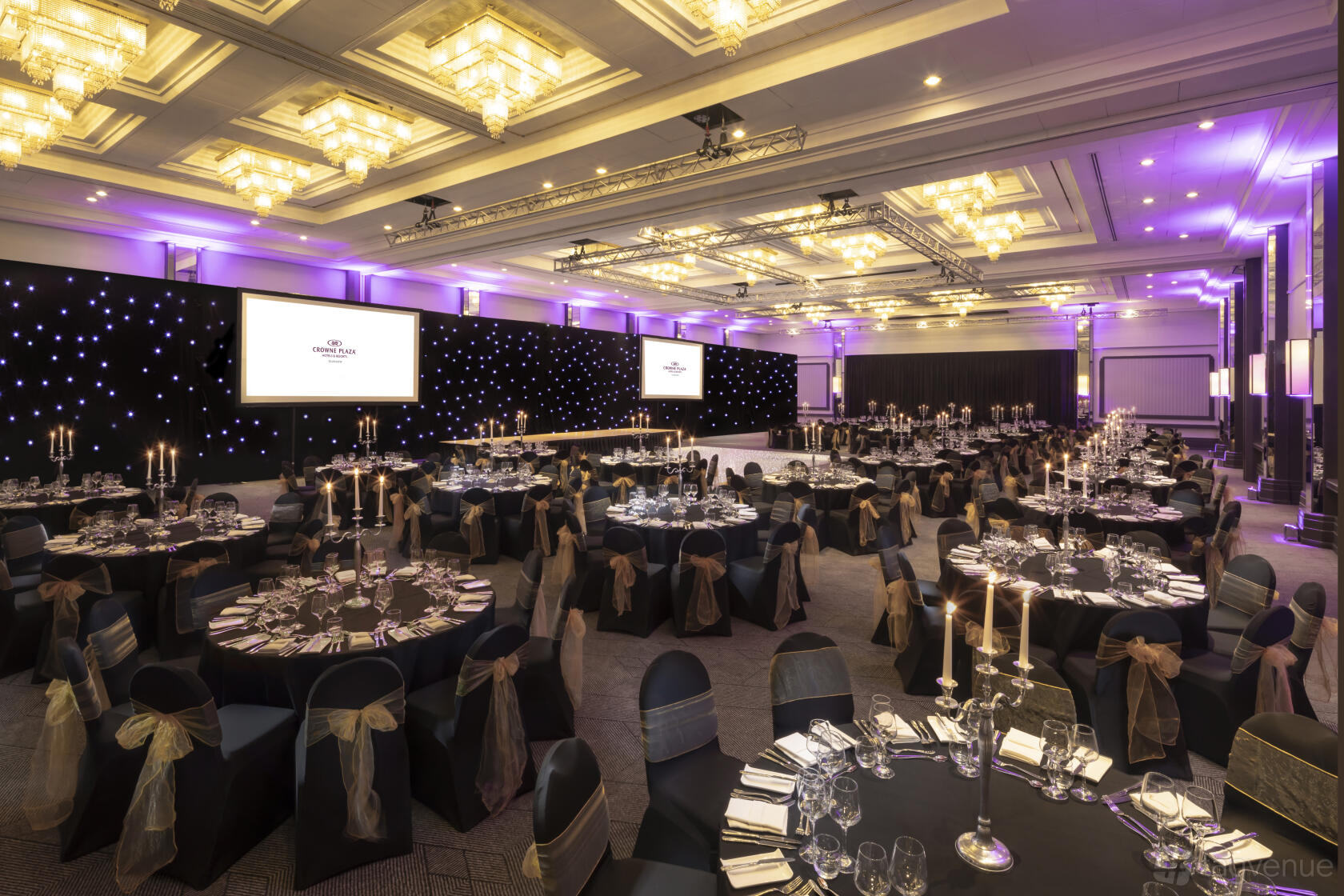 A banqueting hall with round tables, gold sashed chairs, candelabras, and stage lighting at Crowne Plaza Glasgow Argyll Suite.