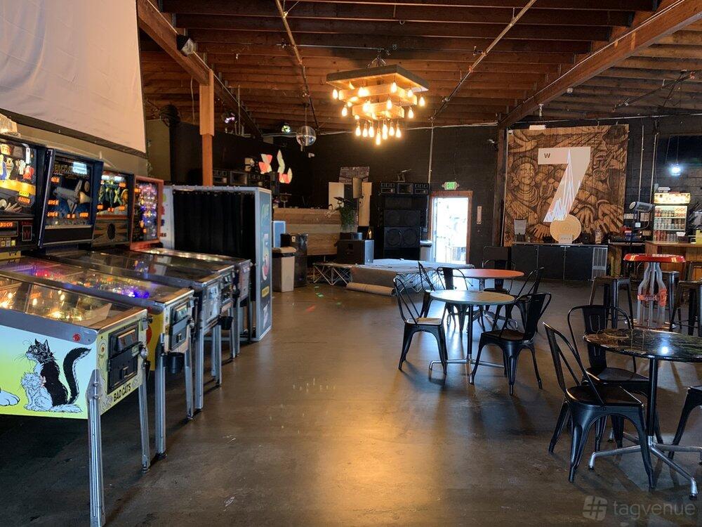A bar with pinball machines, exposed wooden rafters, metal chairs, and a mural at 7th West.