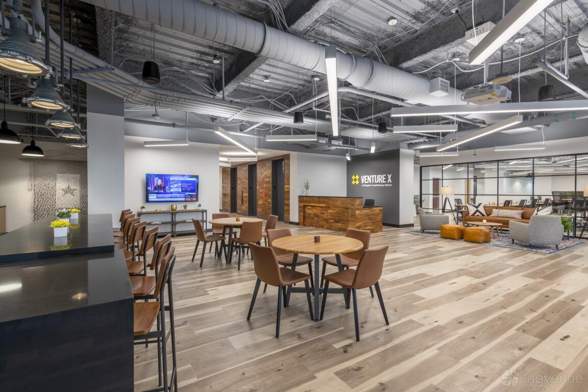 An event space with round wooden tables, brown chairs, exposed ductwork, and a reception desk at Venture X Arlington - Courthouse Metro.