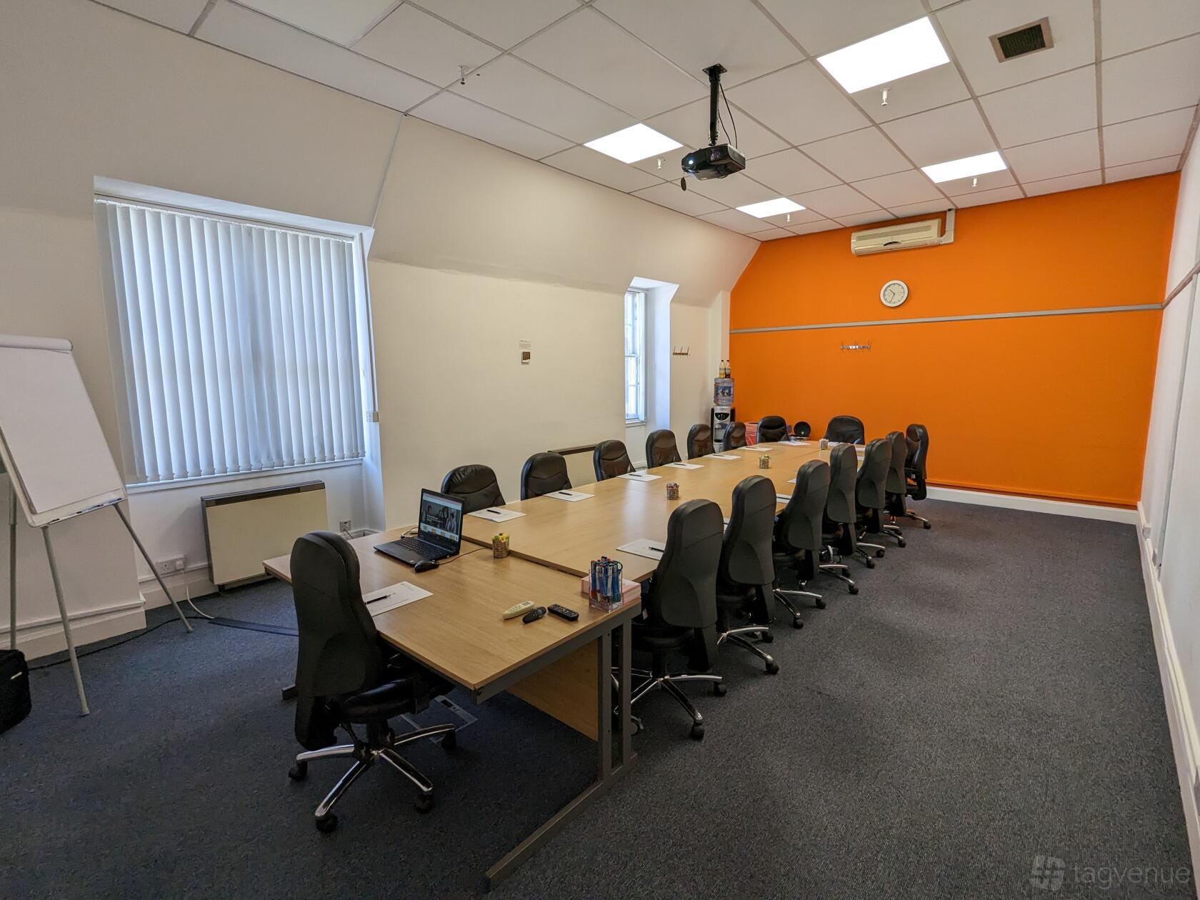 A meeting centre room with a long boardroom table, black chairs, projector, flipchart, and orange accent wall at Edinburgh Training and Conference Venue.