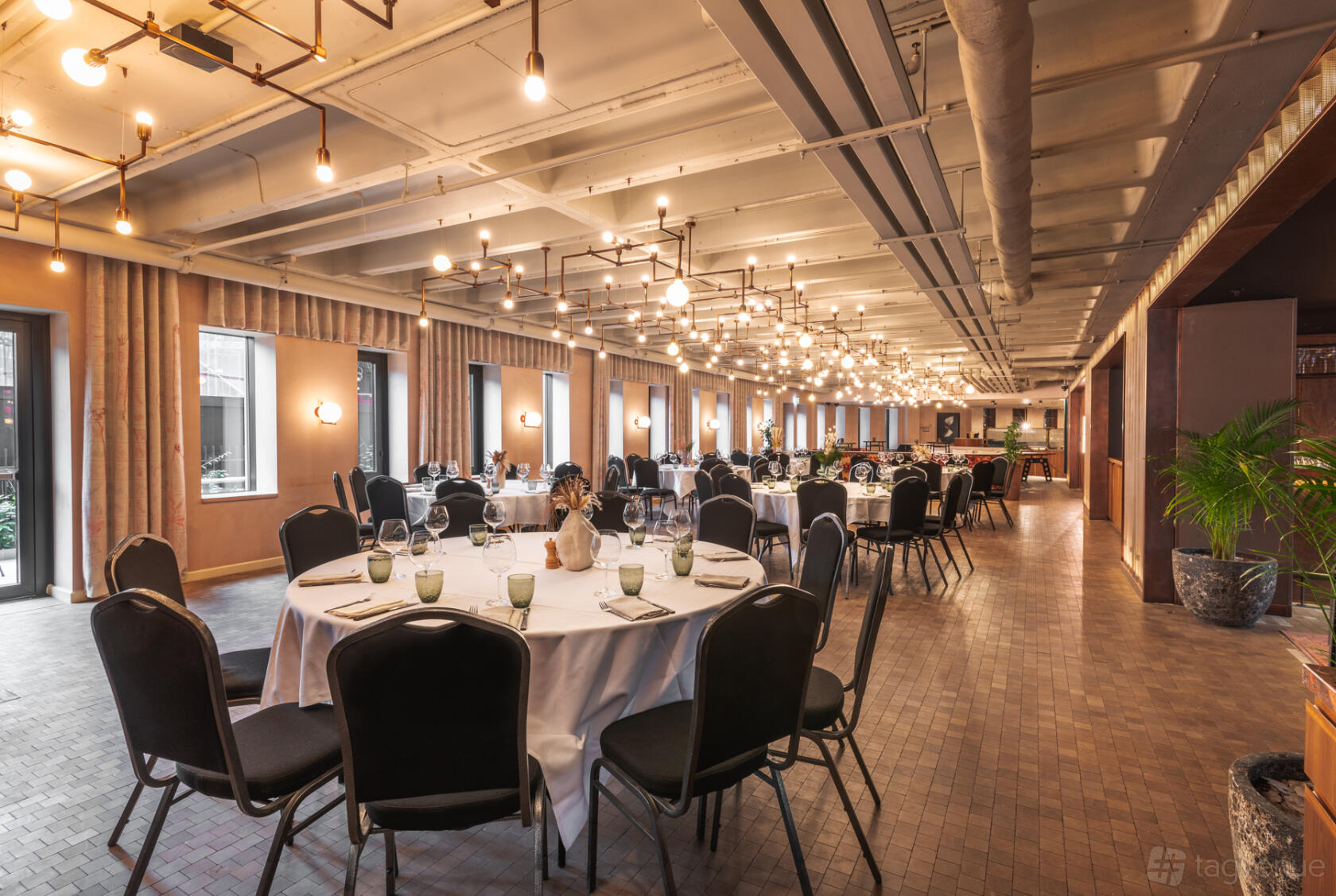 A hotel event space with round tables covered in white linens, black chairs, and hanging pendant lighting at Canopy by Hilton London City.