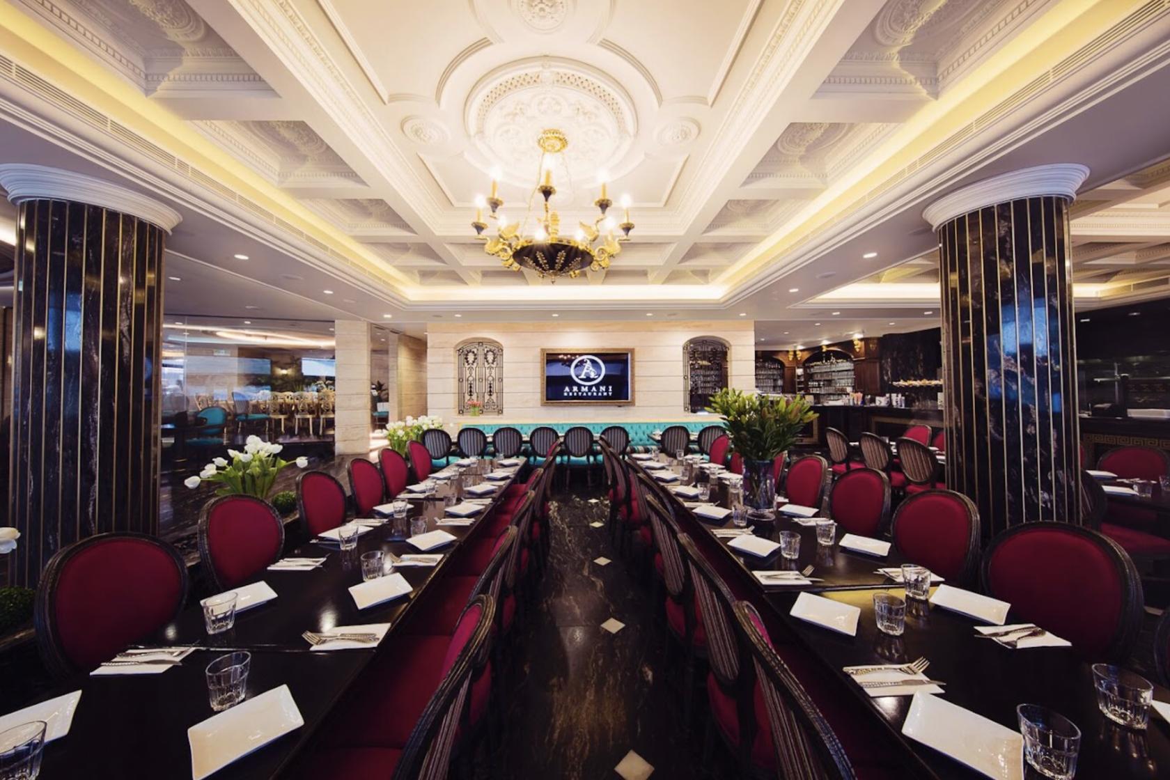 A formal dining room with ornate ceiling moldings, chandeliers, and long tables with red chairs at Armani Restaurant.
