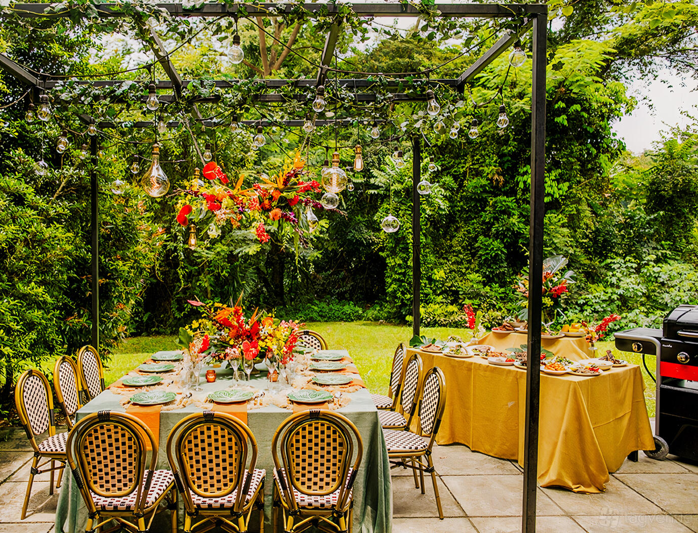 A covered terrace with a long table set for dining, surrounded by greenery and hanging glass lights at Siri House.