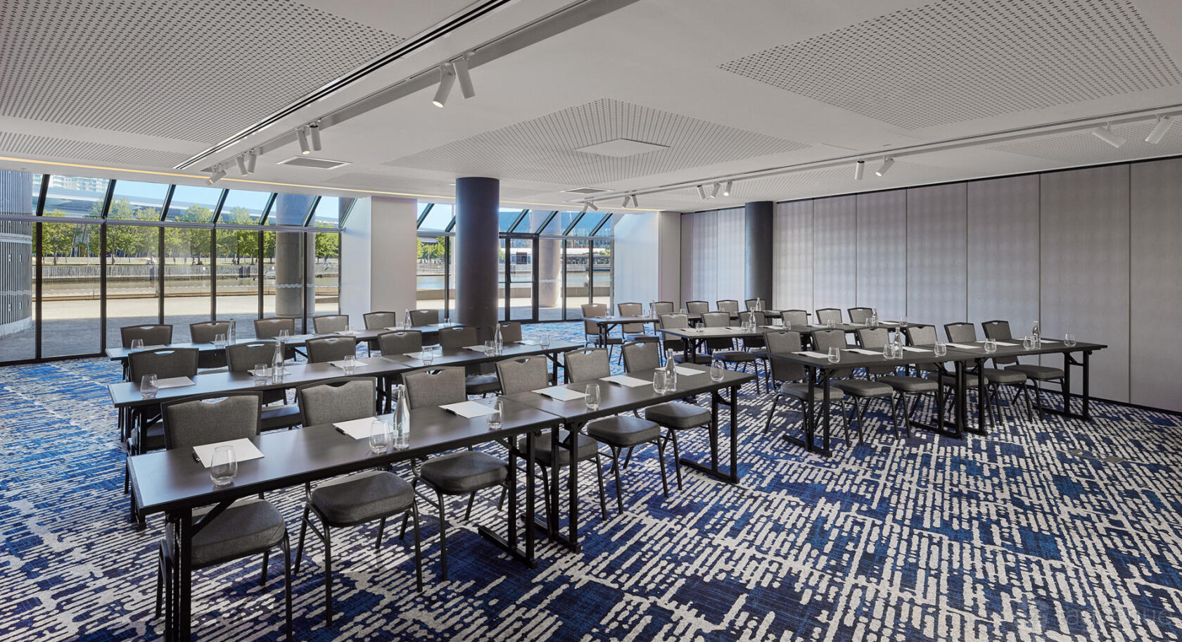 A meeting room with floor-to-ceiling windows, natural lighting, and rows of tables with notepads at Pearl Riverfront.