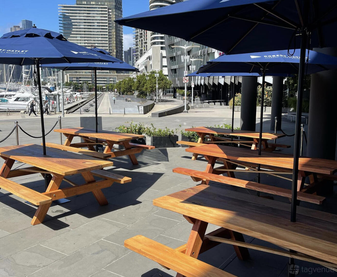 A restaurant terrace with wooden picnic tables and blue umbrellas overlooking a marina at The Espressonist.